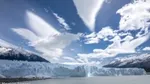 View of the terminus of a glacier as seen from sea level with mountains in the background and a bright, sunny sky